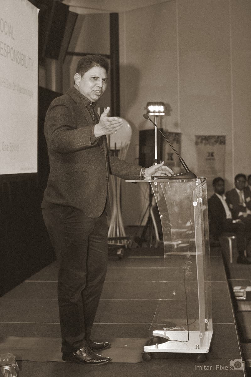 A man in a dark blazer speaks at an OMPEG lectern, one arm extended in an open gesture.