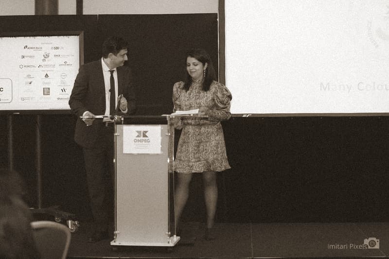 A man and a woman share a lectern on stage at the OMPEG 5th anniversary, mid-conversation, an event screen behind them.