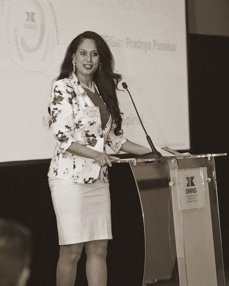 A woman in a floral jacket speaks at an OMPEG lectern, mid-word, one hand resting on her notes.