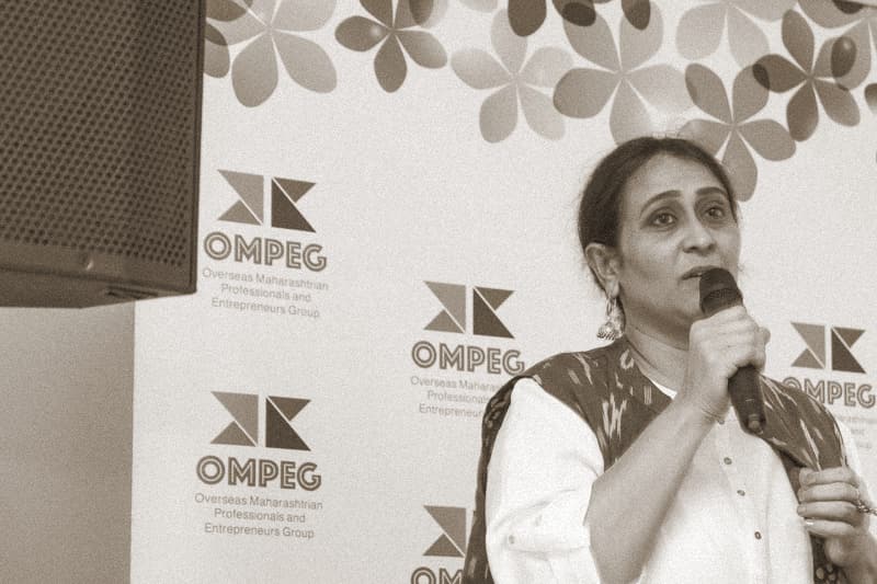 A woman in a white kurta and a pink ikat dupatta speaks into a microphone at an OMPEG Metro Reading event, an orange laptop open on the stand beside her.