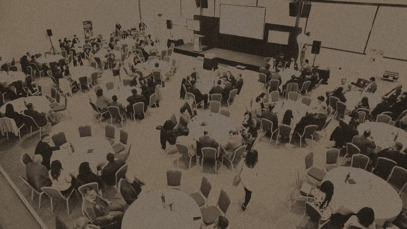 An elevated wide view over the whole banquet hall at the OMPEG 5th anniversary: dozens of round tables, colourful uplighting, a stage with a candle graphic on the screen, no single person dominant in frame.