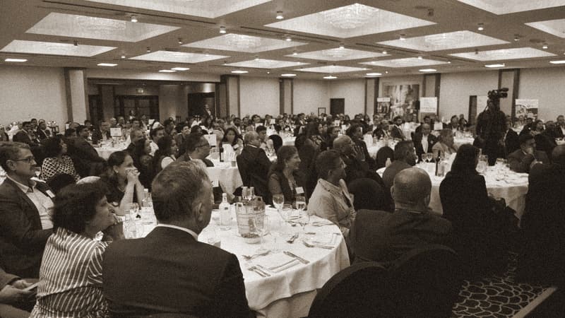 A packed banquet hall seen from the back, a close foreground pair with their backs to camera anchoring the bottom of the frame, a distant stage barely visible top-left.